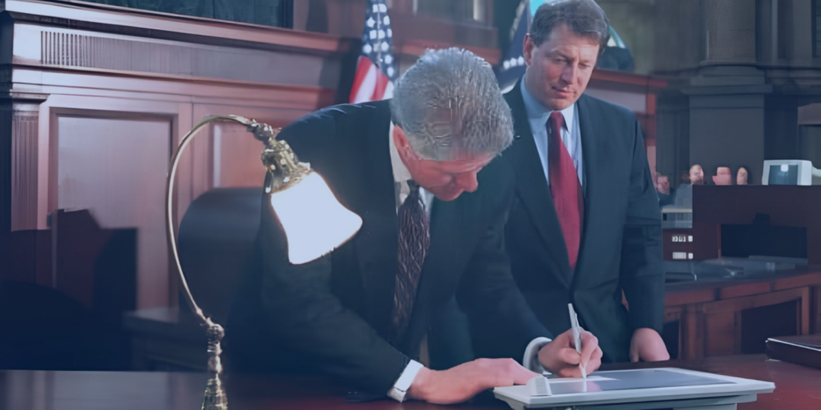 President Bill Clinton signs a document in a formal government setting while Al Gore stands behind him watching. An American flag and wood-paneled interior are visible in the background.